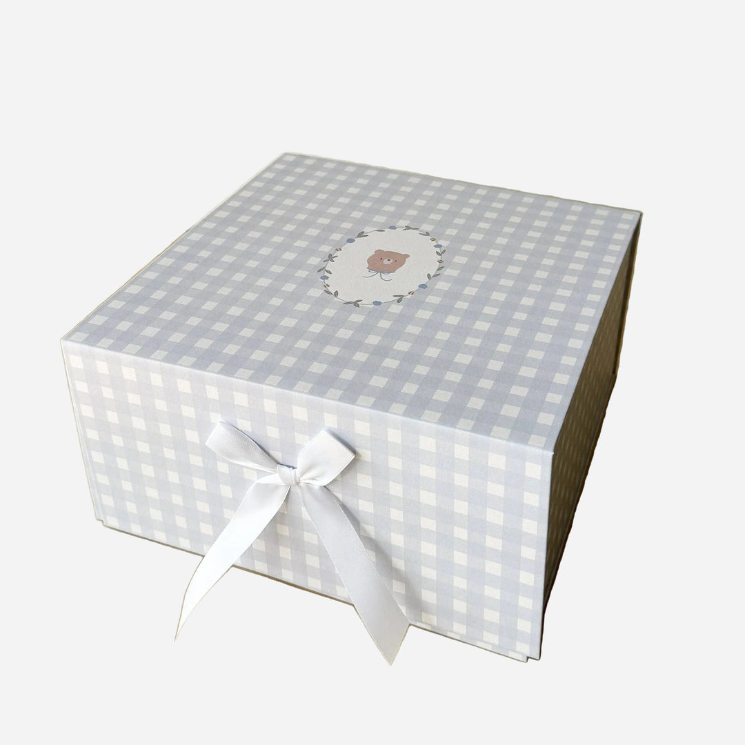 A square gift box with a light blue and white gingham pattern, tied with a white ribbon bow on the front. The top lid features a small circular illustration of a teddy bear in a floral frame.
