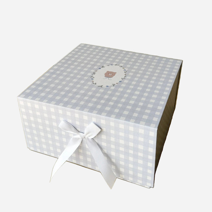 A square gift box with a light blue and white gingham pattern, tied with a white ribbon bow on the front. The top lid features a small circular illustration of a teddy bear in a floral frame.