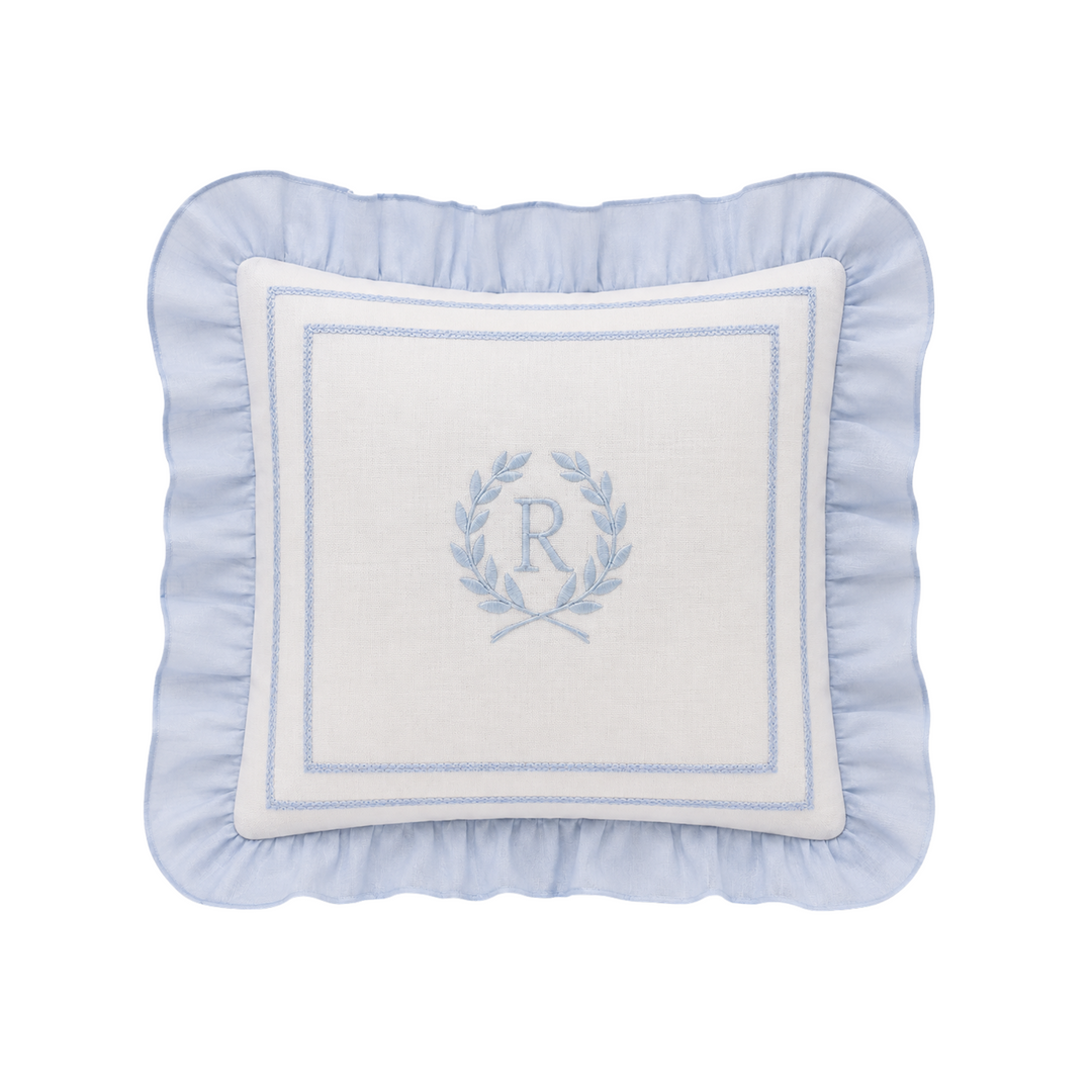 A square decorative throw pillow with a light blue ruffled border and a white center panel featuring a light blue embroidered monogram wreath with the letter 'R'.