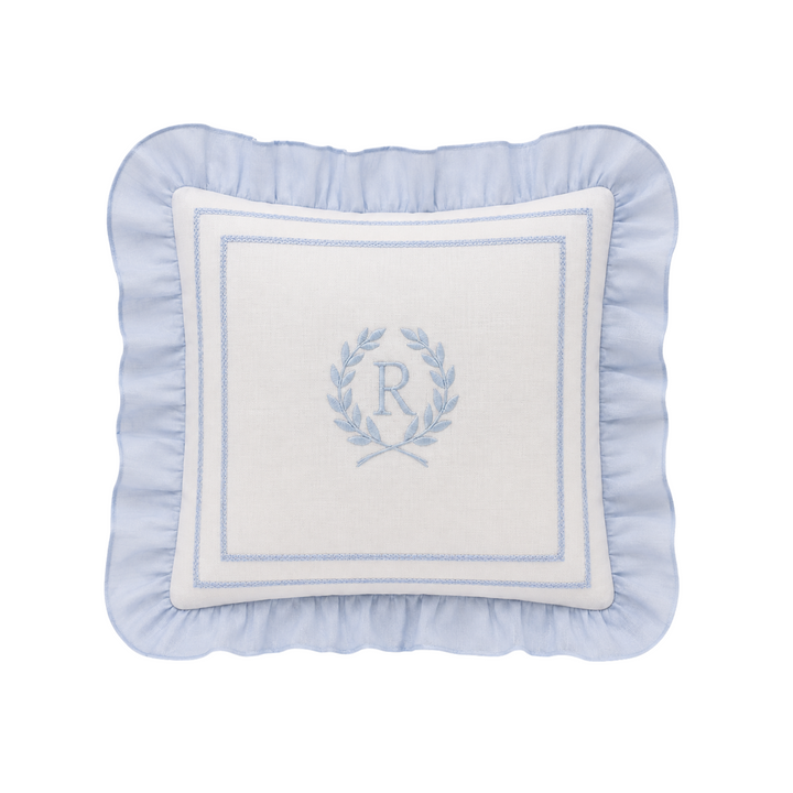 A square decorative throw pillow with a light blue ruffled border and a white center panel featuring a light blue embroidered monogram wreath with the letter 'R'.