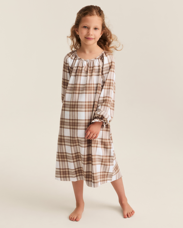 A young girl with curly light brown hair stands barefoot against a plain light beige studio background, wearing a long-sleeve nightgown with a brown and white plaid pattern.