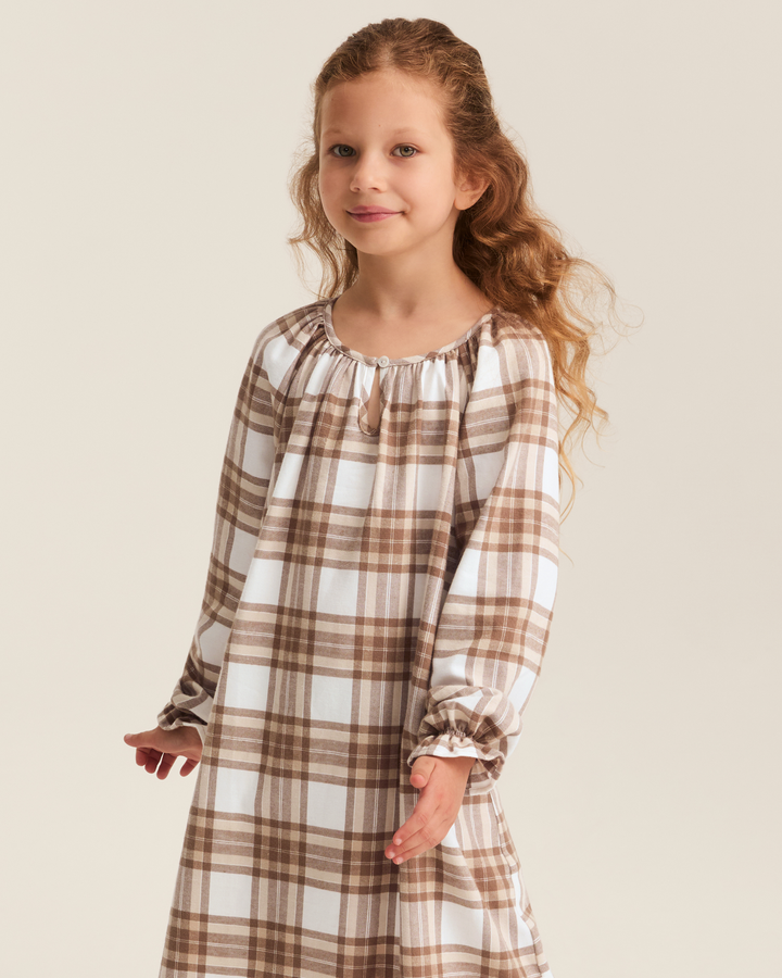 A young girl with long wavy light brown hair wearing a long-sleeved plaid nightgown in brown and white, standing against a solid light beige background.