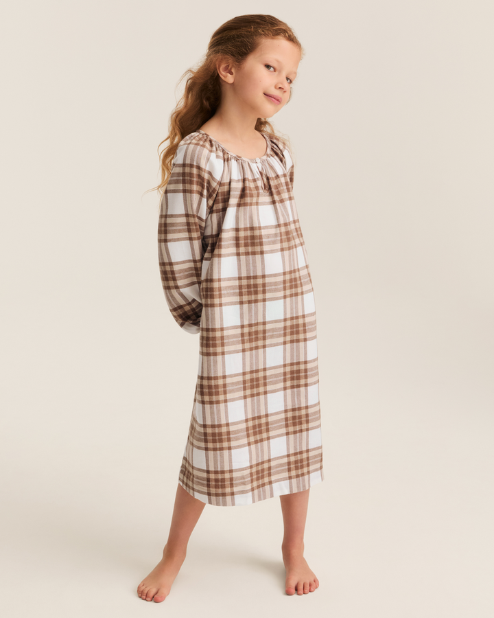 A young girl with long light brown hair stands barefoot against a warm beige studio background, wearing a long-sleeve nightgown with a brown and white plaid pattern.