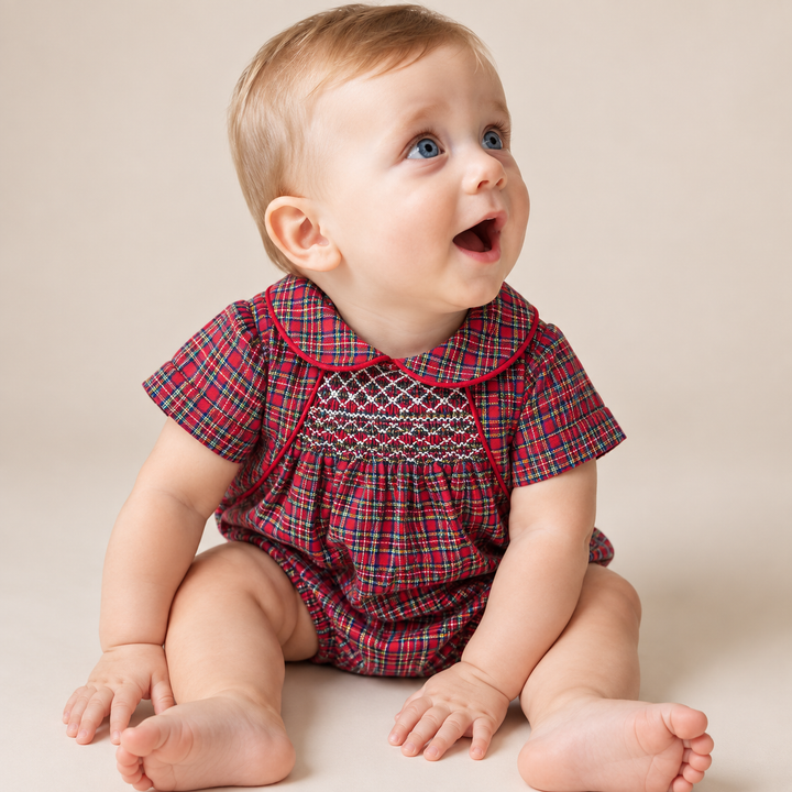 A baby sitting on a beige studio floor wearing a red and green plaid short-sleeve romper with white smocked detailing on the chest.