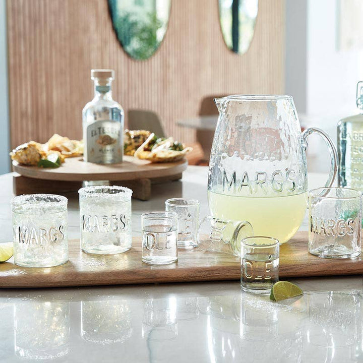 Margarita glasses and pitcher on a table with snacks and bottles in the background