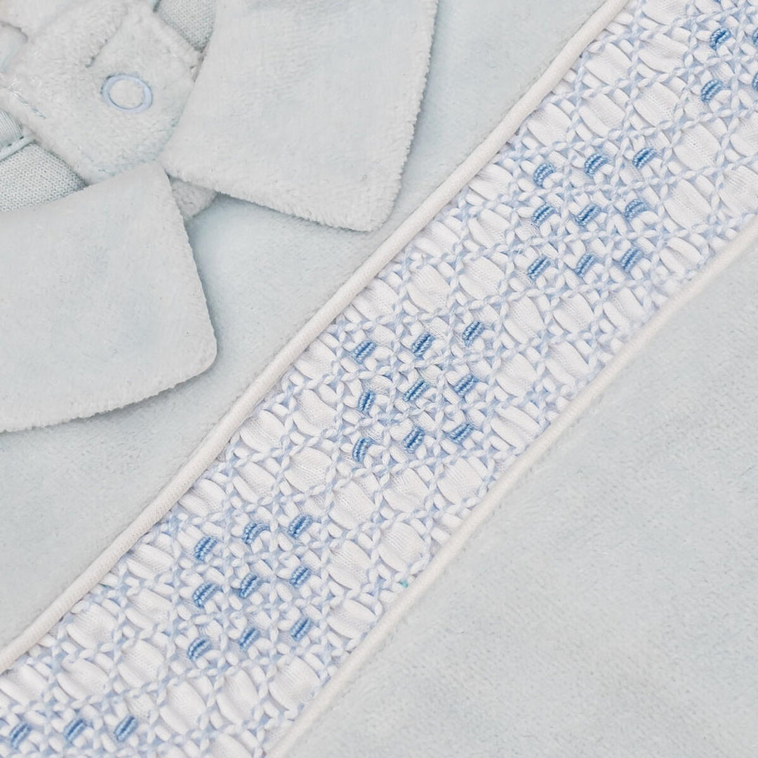 Close-up of a light blue plush fabric item, likely a soft accessory or garment, featuring a decorative white and light blue embroidered border with a small button detail.