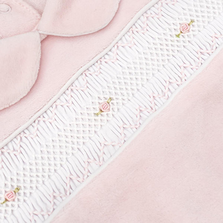 Close-up of a baby garment or textile detail in soft pink and white, featuring a smocked white panel with small embroidered pink rose motifs and a curved pink fabric section at the top.