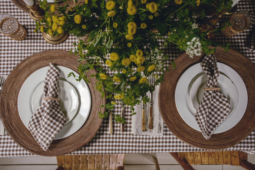 A styled dining table setting viewed from above, featuring two matching place settings with white plates, round woven brown placemats, brown-and-white gingham napkins tied with string, a brown-and-white gingham tablecloth, and a large yellow flower centerpiece.