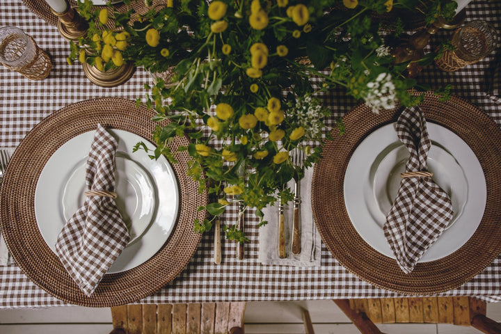 A styled dining table setting viewed from above, featuring two matching place settings with white plates, round woven brown placemats, brown-and-white gingham napkins tied with string, a brown-and-white gingham tablecloth, and a large yellow flower centerpiece.