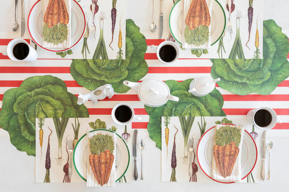 Table setting with vegetable-themed placemats, plates, and cutlery on a striped tablecloth.