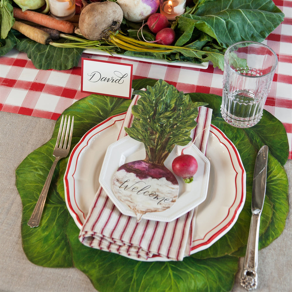 Table setting with green leafy vegetables, a red and white checkered tablecloth, and a name card.