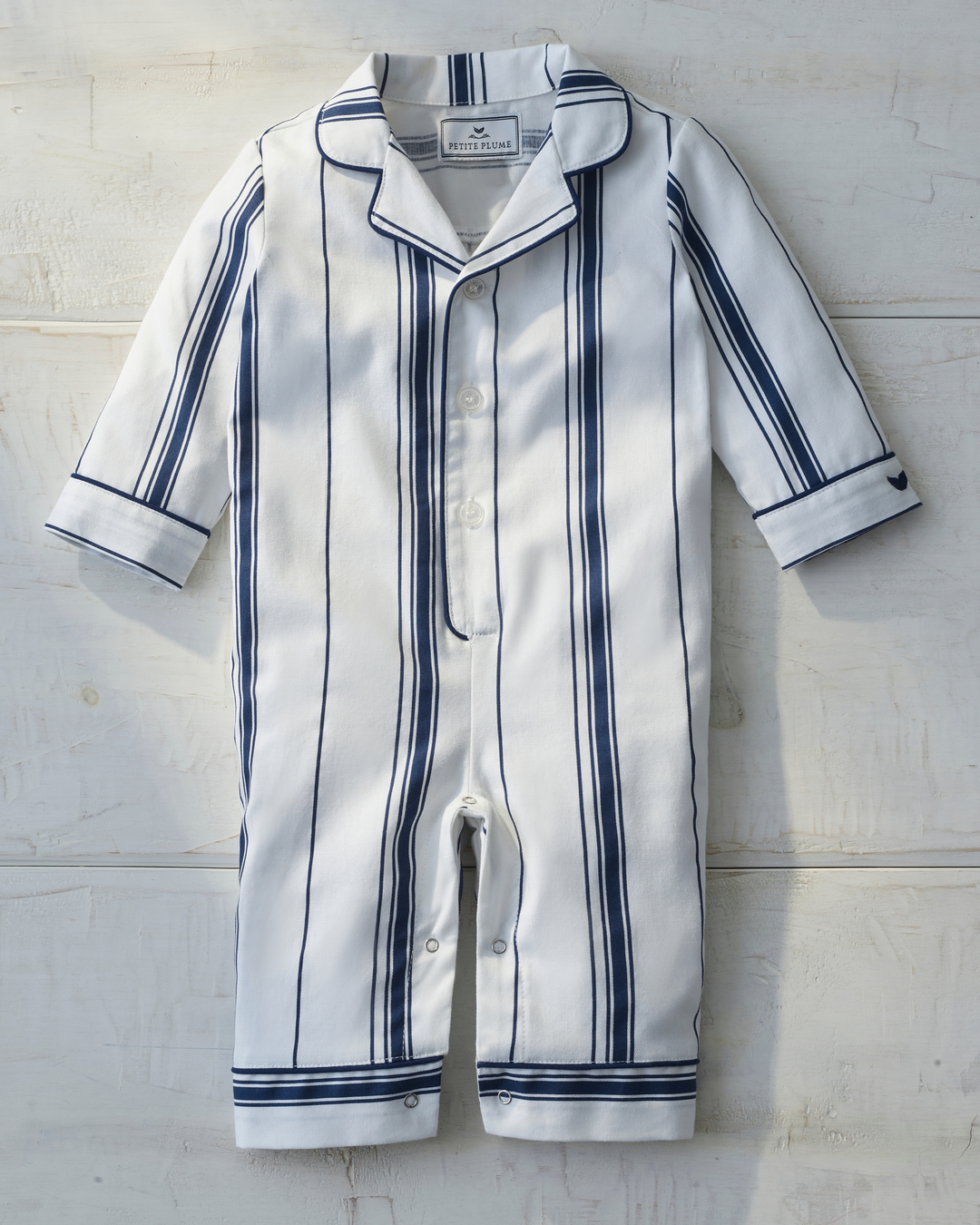 A baby romper in white fabric with navy blue vertical pinstripes, featuring a collar, short sleeves, and snap closures, laid flat on a white wooden background.