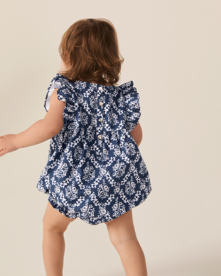 A toddler girl seen from behind wearing a navy blue and white floral print romper with ruffled shoulder straps, set against a plain beige studio background.