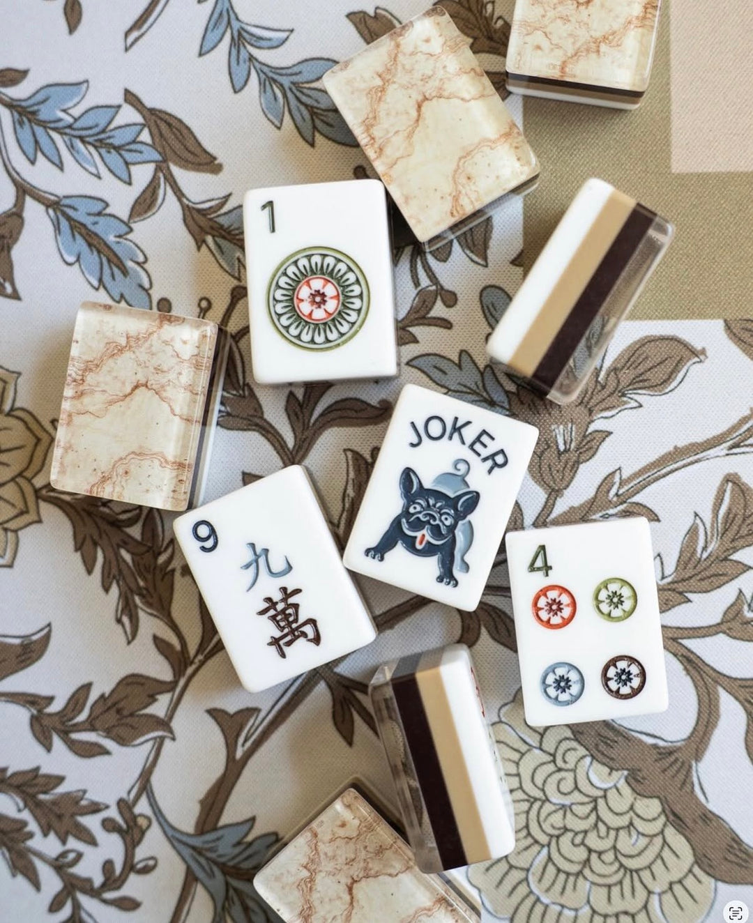A set of Mahjong tiles arranged on a decorative floral-patterned surface. The tiles include cream and beige marble-look backs, a white Joker tile with a black dog illustration, and other visible numbered tiles with Chinese characters and colored symbols.