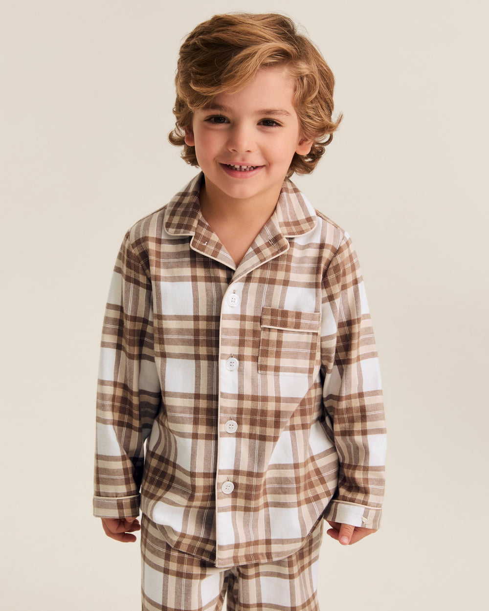 A young boy with wavy light brown hair smiling, wearing a two-piece plaid pajama set in brown, beige, and white. The set includes a long-sleeve button-up shirt and matching pants, photographed against a solid light beige studio background.