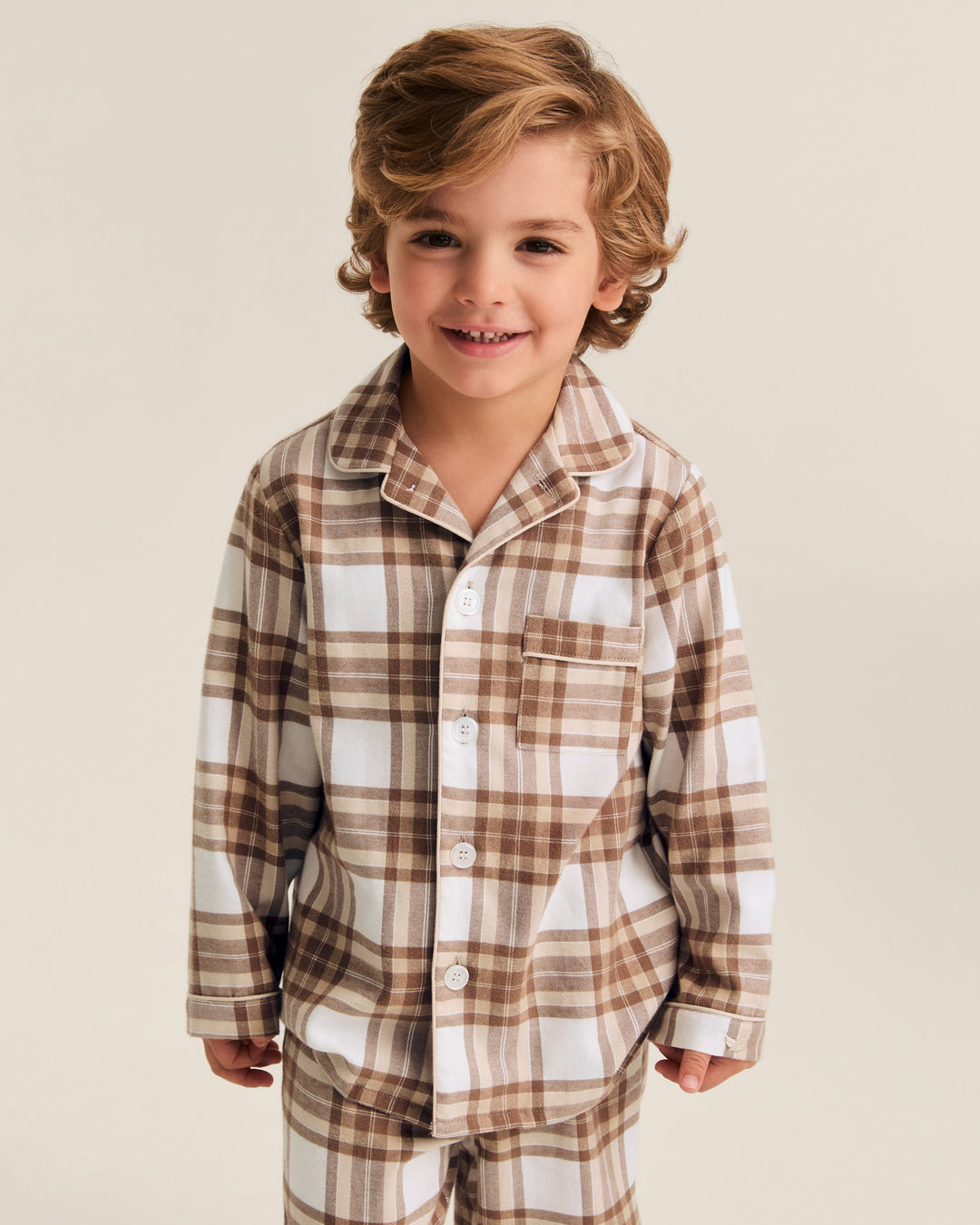 A young boy with wavy light brown hair smiling, wearing a two-piece plaid pajama set in brown, beige, and white. The set includes a long-sleeve button-up shirt and matching pants, photographed against a solid light beige studio background.