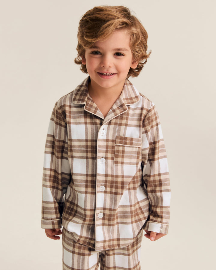 A young boy with wavy light brown hair smiling, wearing a two-piece plaid pajama set in brown, beige, and white. The set includes a long-sleeve button-up shirt and matching pants, photographed against a solid light beige studio background.