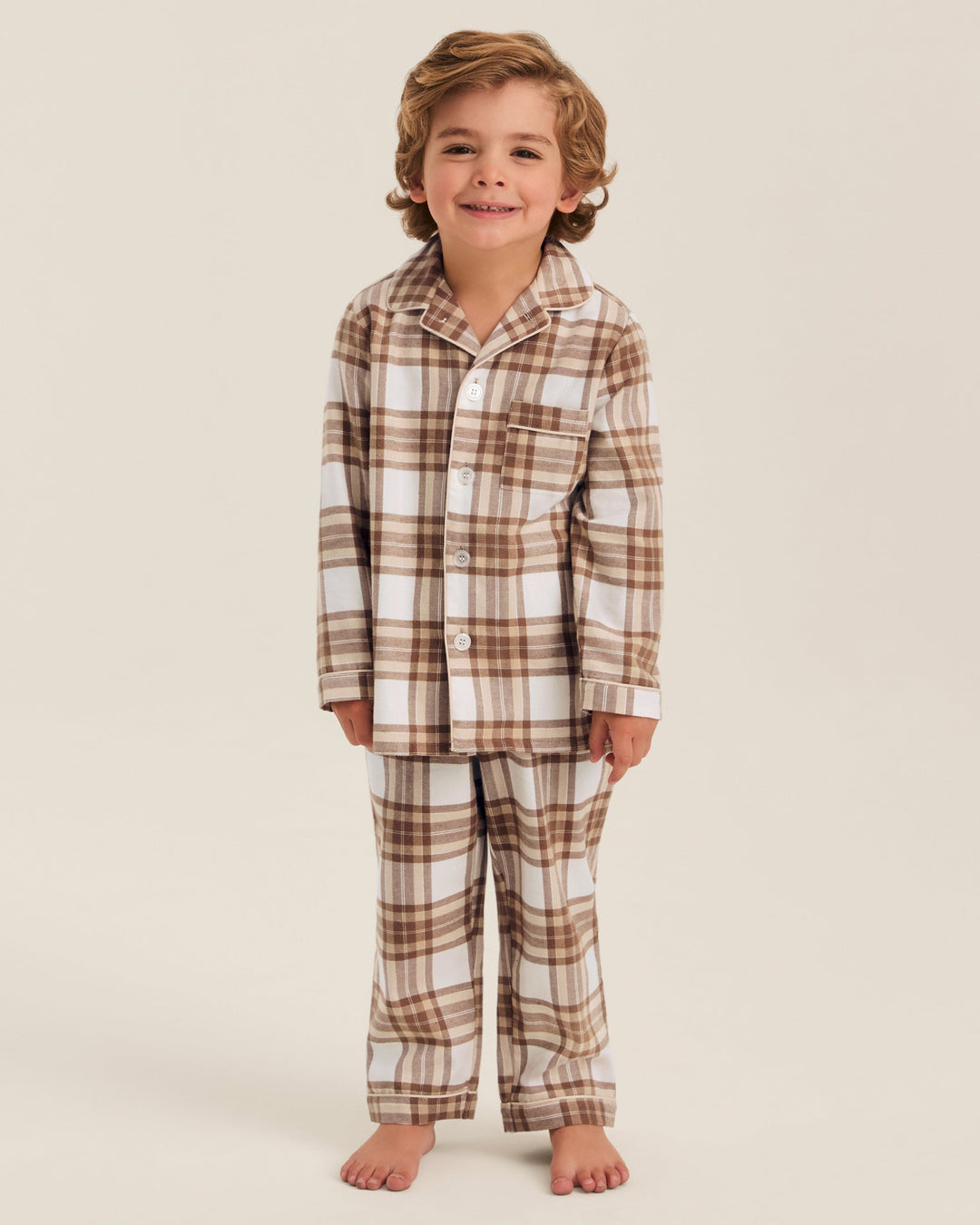 A young child wearing a two-piece pajama set with a brown and beige plaid pattern, standing against a solid light beige background.