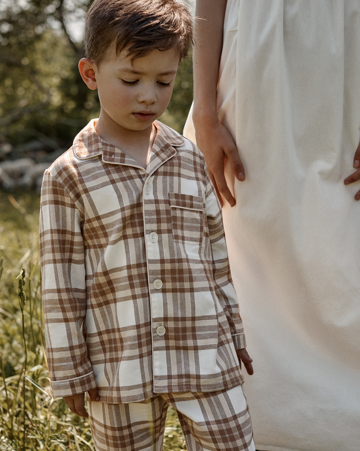 A young boy wearing a brown and beige plaid pajama set stands outdoors next to an adult in a white dress, with a soft-focus natural background.