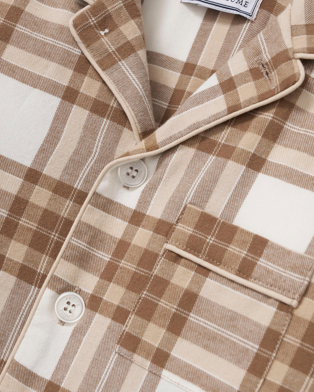 Close-up of a plaid button-front shirt or pajama top in warm beige, tan, and brown tones, with a collar, white buttons, and a chest pocket. A small brand tag is partially visible at the collar.