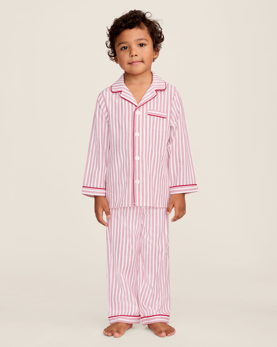 A young child wearing a red and white vertically striped pajama set with solid pink piping, standing against a plain light beige studio background.