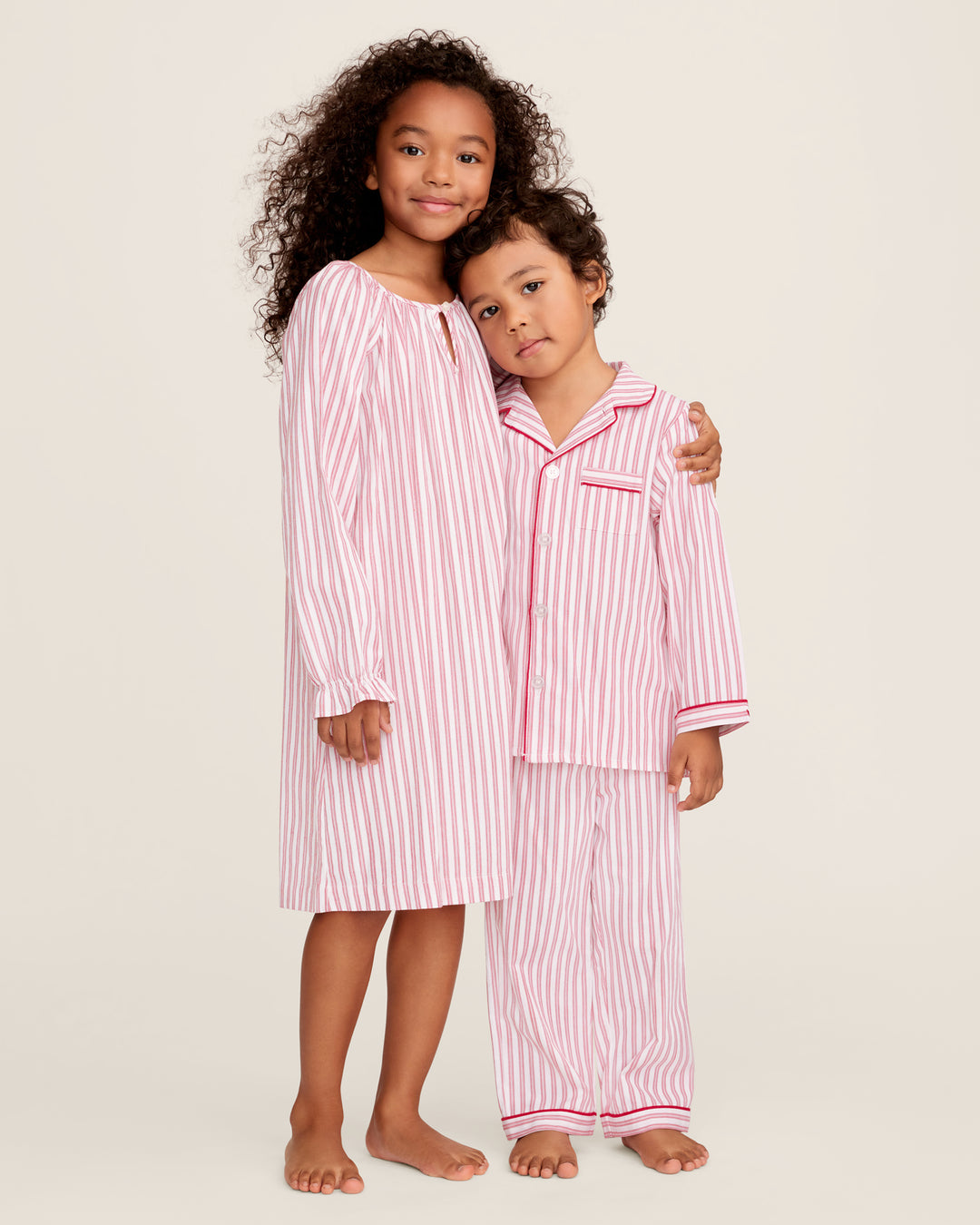 Two children modeling matching red and white striped pajamas against a plain beige studio background. The taller child wears a long-sleeve pajama top and pants set, and the shorter child wears a short-sleeve pajama top with matching pants.