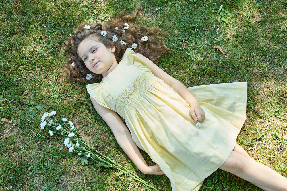 Young girl in a yellow dress lying on grass with flowers in her hair and around her.