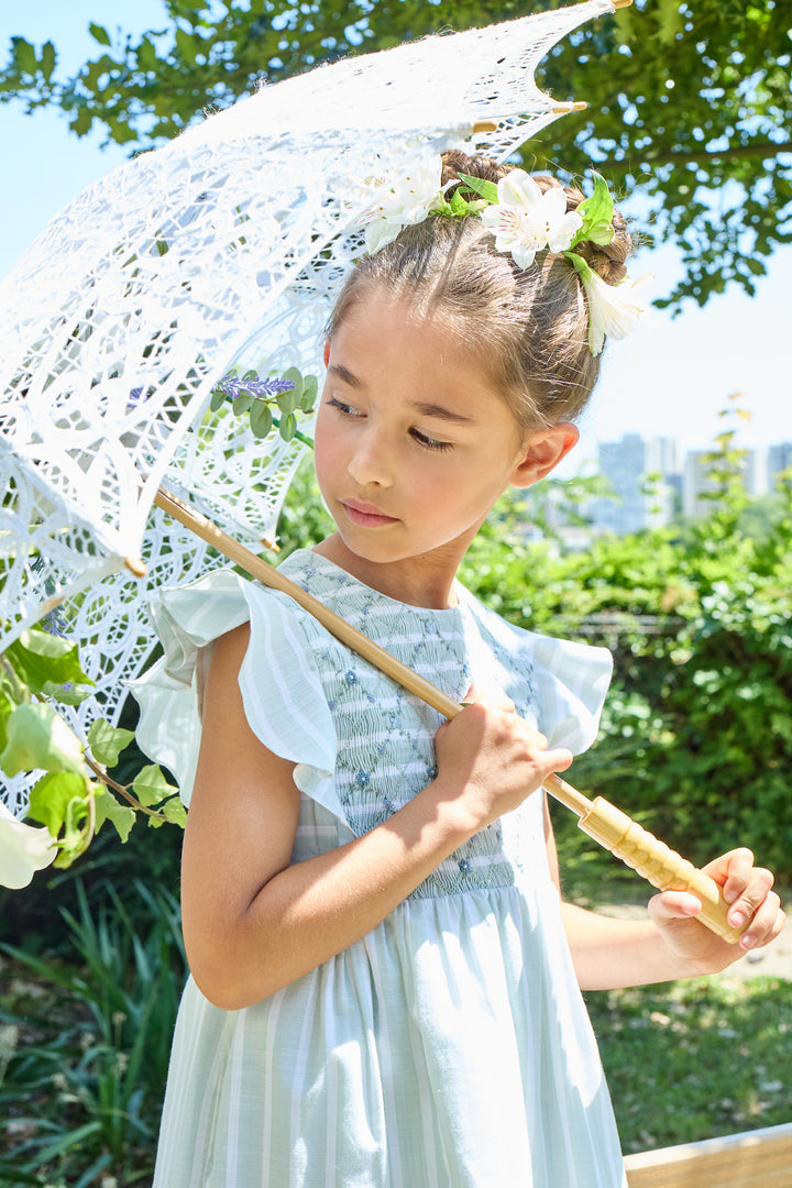Young girl in a light dress holding a lace parasol outdoors with greenery and a cityscape in the background.