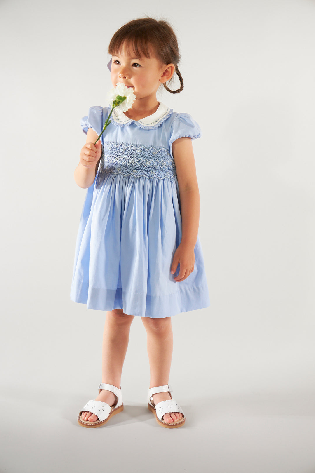 Young girl wearing a light blue dress with lace details, holding a flower against a plain background