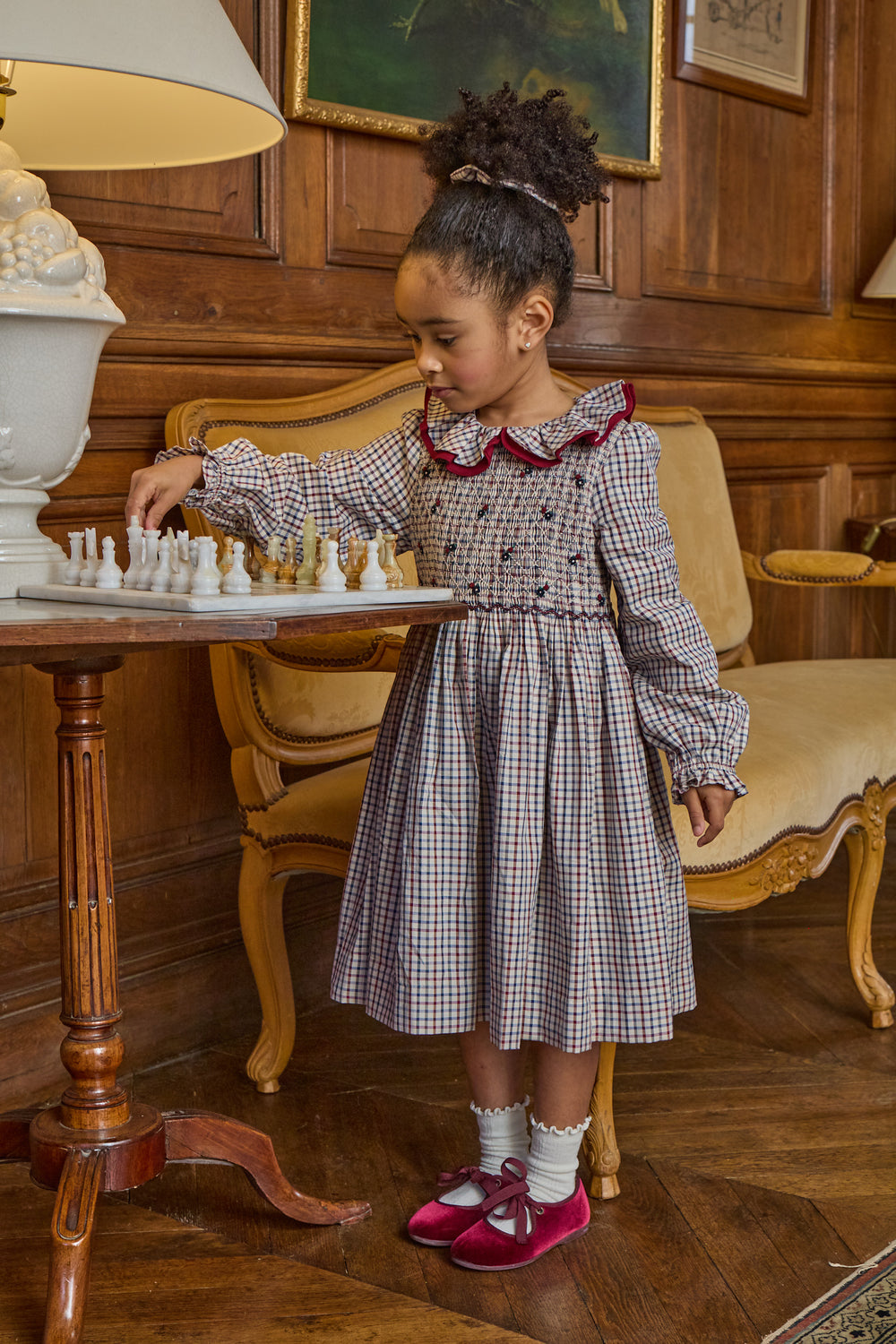 A young girl with curly hair stands in a vintage room next to a wooden table, wearing a long-sleeved gingham dress with a dark red collar trim and pink shoes. She is looking down at a chessboard on the table.