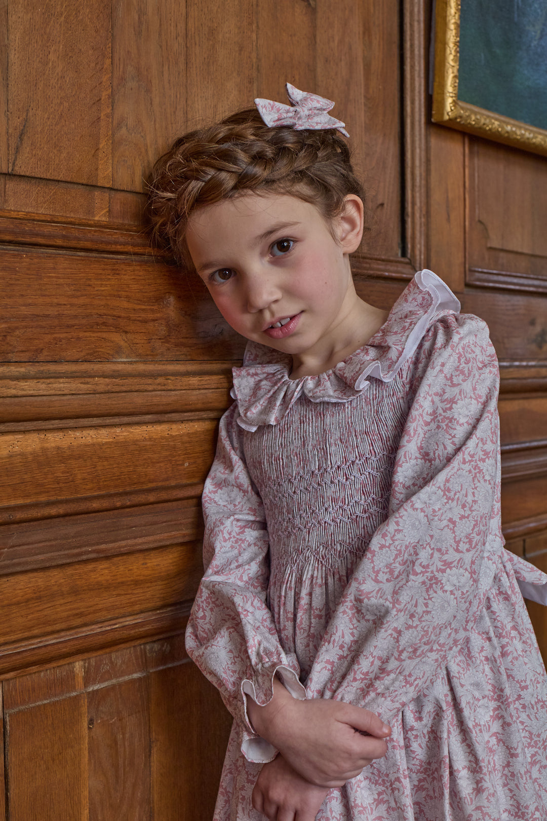 A young girl with braided hair and a pink bow accessory, wearing a smocked long-sleeve dress with a pink and white floral pattern, leaning against a wooden paneled wall.