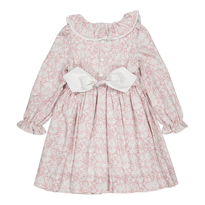 A long-sleeve baby girl's dress with a pink and white floral toile pattern, featuring a large white bow at the waist, a white ruffled collar, and buttoned cuffs.