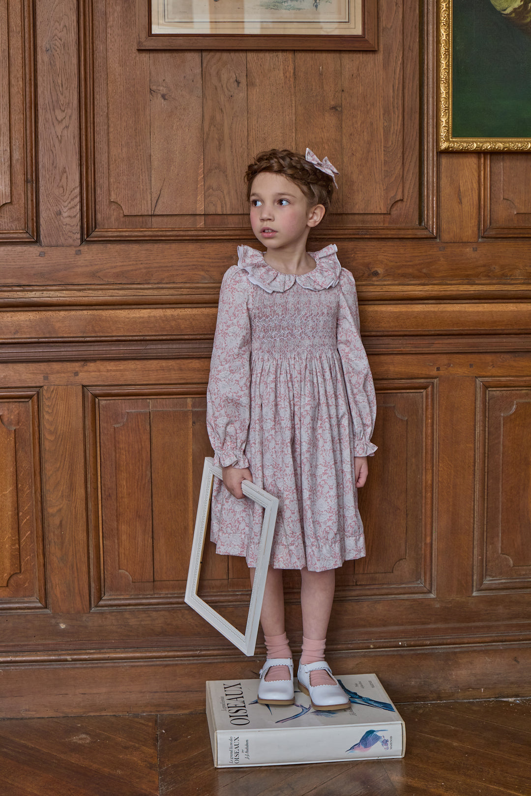A young girl stands on a book in front of a wooden paneled wall, wearing a light pink and white floral smocked dress with ruffled shoulders and holding a white picture frame.