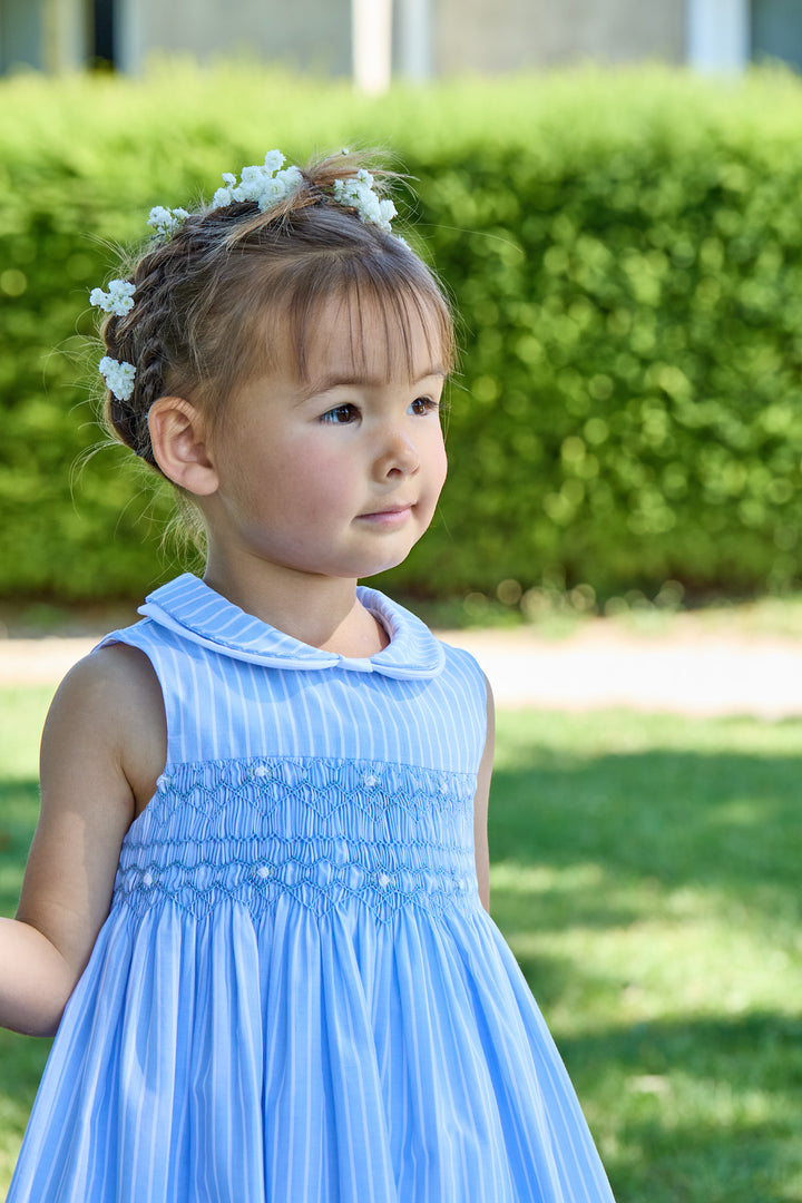 Young girl in a light blue dress with braided hair and flower headband standing outdoors.