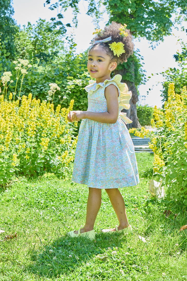 Young girl in a floral dress standing in a garden with yellow flowers and greenery.