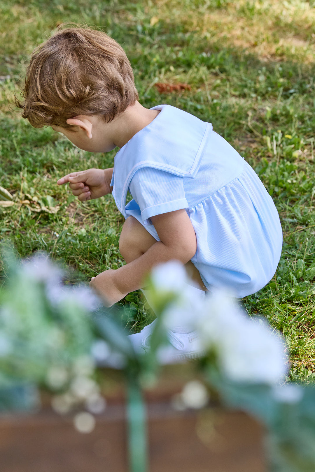 Child in a light blue outfit sitting on grass with flowers in the foreground