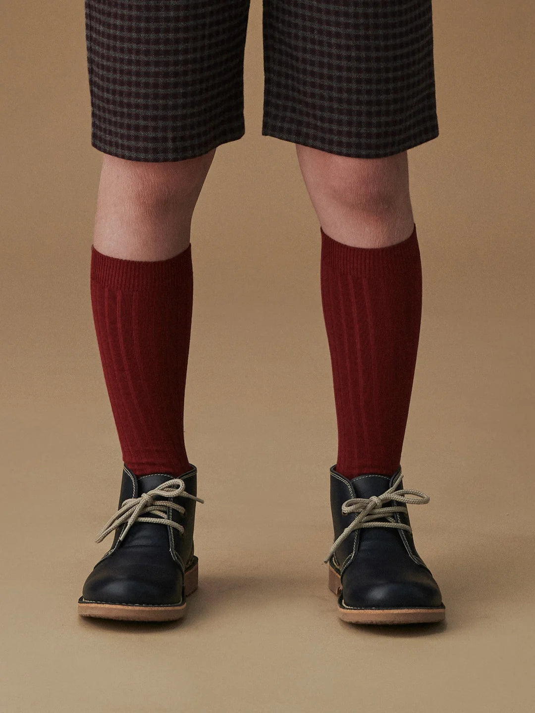 Person wearing dark checkered shorts, red knee-high socks, and black shoes on a brown background