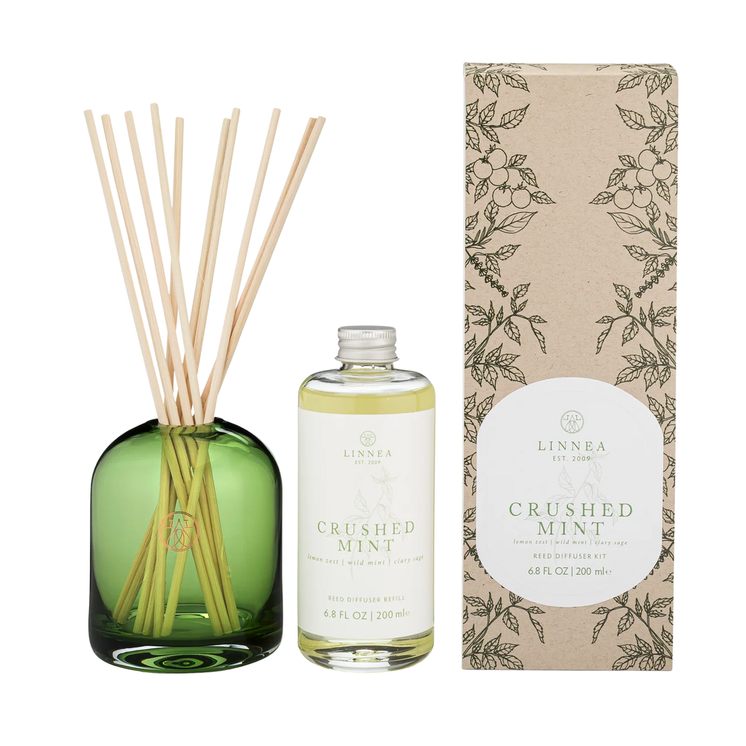 LINNEA Crushed Mint reed diffuser set featuring a green glass bottle with natural reed sticks, a clear glass bottle of oil and label, and a brown kraft paper box with botanical illustrations.