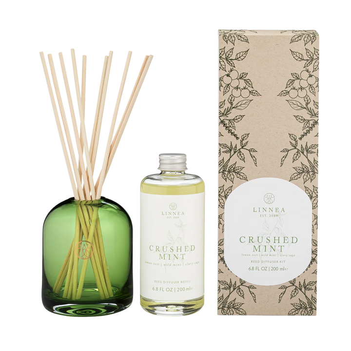 LINNEA Crushed Mint reed diffuser set featuring a green glass bottle with natural reed sticks, a clear glass bottle of oil and label, and a brown kraft paper box with botanical illustrations.