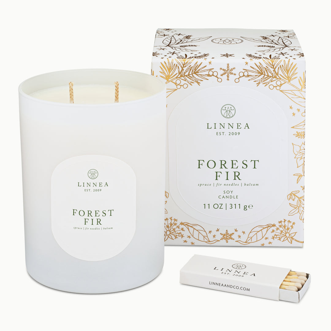 A white soy candle in a matte white glass jar with a round white label reading 'LINNEA EST. 2009 FOREST FIR', shown next to its matching white and gold floral-patterned box and a small white matchbox on a clean white background.