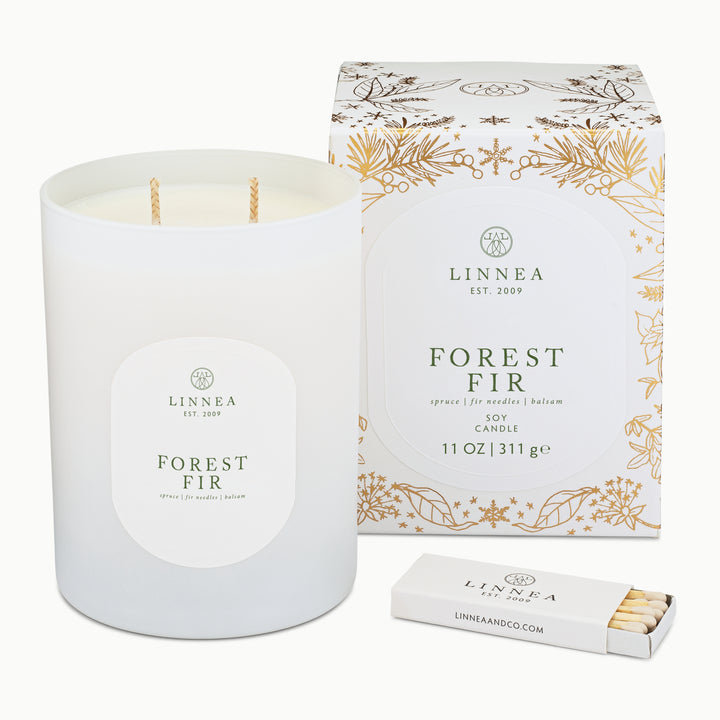 A white soy candle in a matte white glass jar with a round white label reading 'LINNEA EST. 2009 FOREST FIR', shown next to its matching white and gold floral-patterned box and a small white matchbox on a clean white background.