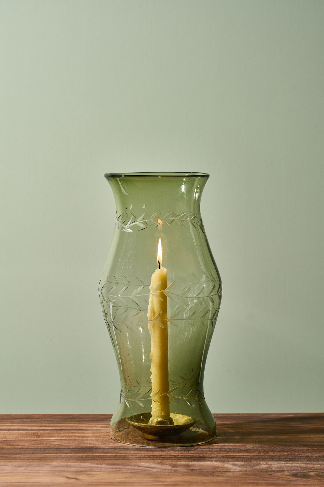 An olive green glass candle holder with etched leaf patterns, containing a lit yellow taper candle, set on a wooden surface against a pale sage green background.