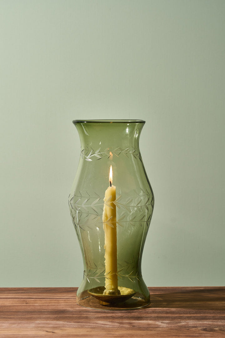 An olive green glass candle holder with etched leaf patterns, containing a lit yellow taper candle, set on a wooden surface against a pale sage green background.