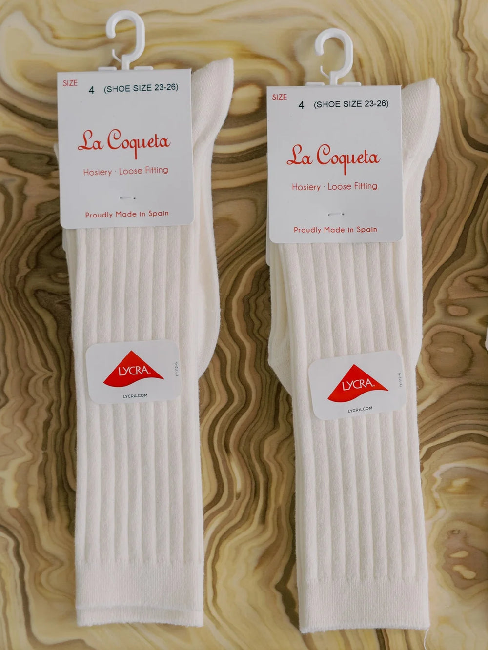 Two pairs of white socks on hangers with 'La Coqueta' labels on a wooden surface.
