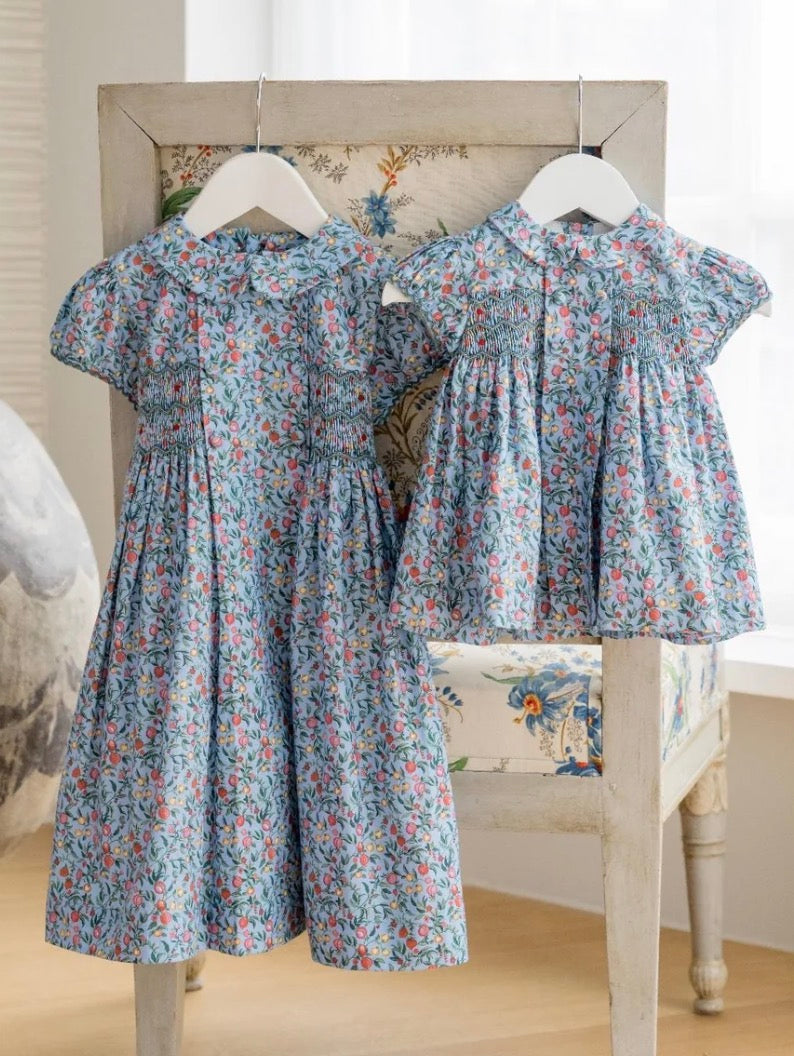 Two girls' short-sleeve floral dresses hanging on white hangers on a rustic wooden chair. The larger dress is blue with red and pink floral print, and the smaller dress is a matching light blue floral print.