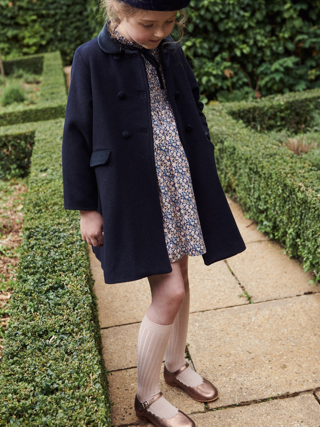 Child wearing a navy coat and floral dress standing in a garden.