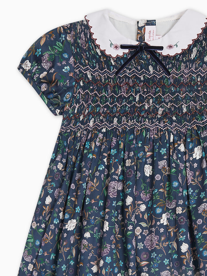 A girls' dress with a navy blue floral print, featuring a white ruffled collar, a dark navy bow at the neckline, and smocked detailing across the chest.