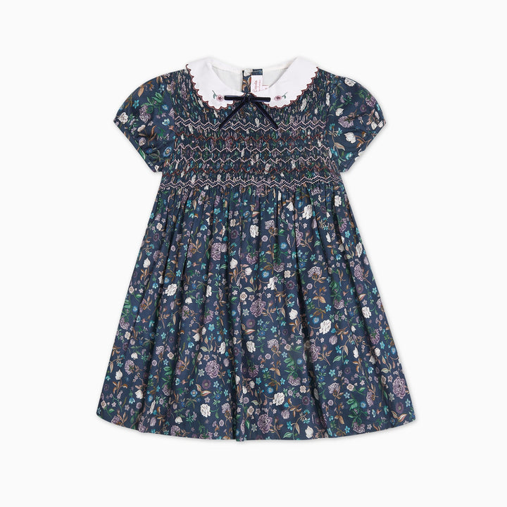 A toddler girl's smocked dress featuring a dark navy blue floral and paisley print, a white ruffled collar, and a black bow at the neckline, shown against a plain white background.
