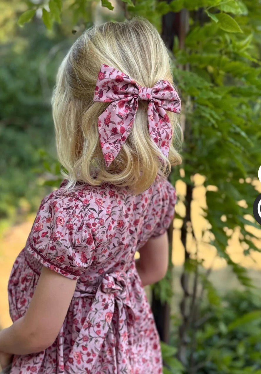 A young girl with blonde hair seen from behind, wearing a pink floral dress and a matching pink floral hair bow, standing outdoors in front of green foliage.