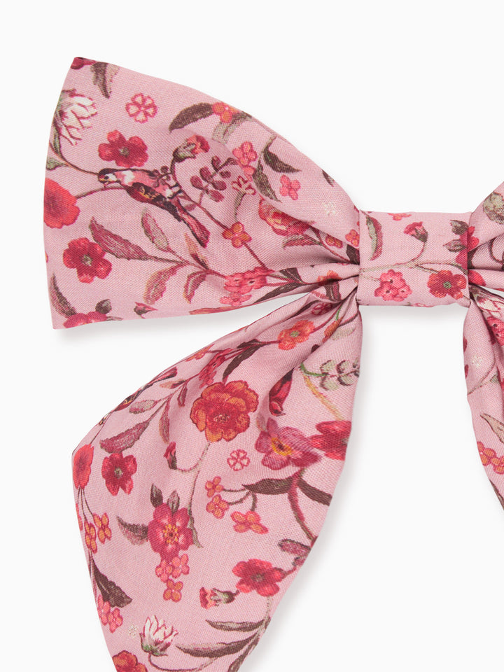A pink bow tie featuring a watercolor-style floral print with red, pink, and brown blossoms and leaves, shown against a plain white background.